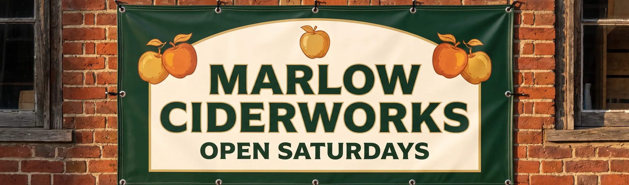 A full-color Marlow Ciderworks vinyl banner stretched across a brick storefront with its stitched hem and metal grommets visible