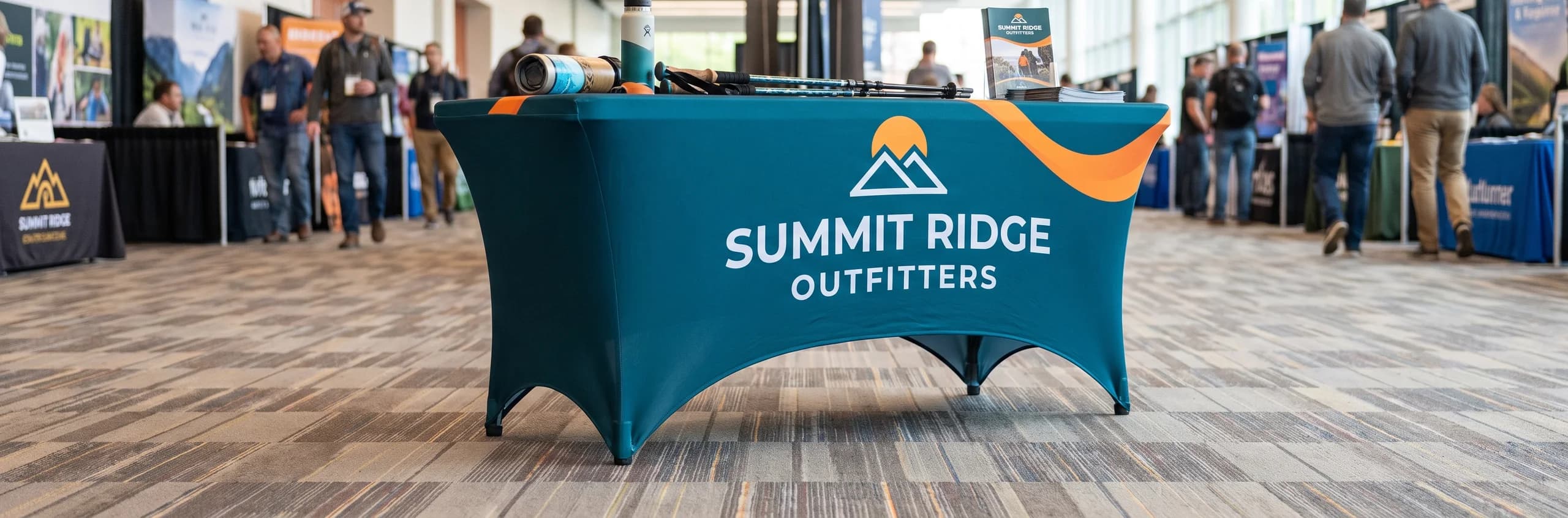 Teal stretch table cover printed with an outdoor outfitter logo and an orange wave, fitted tight over a folding table in a busy expo hall