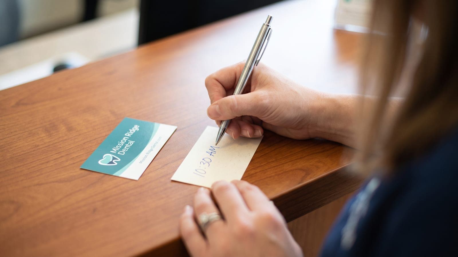 A receptionist writing an appointment time on the back of a Mission Ridge Dental business card at a wooden front desk, the printed front of the same card lying beside it