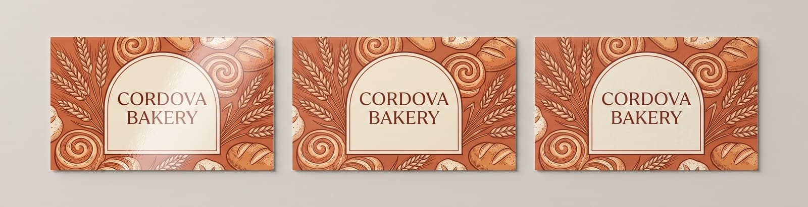 Three full color Cordova Bakery business cards lying in a row under one light, the gloss card reflecting a bright highlight, the matte card flat and even, and the uncoated card soft with a visible paper texture