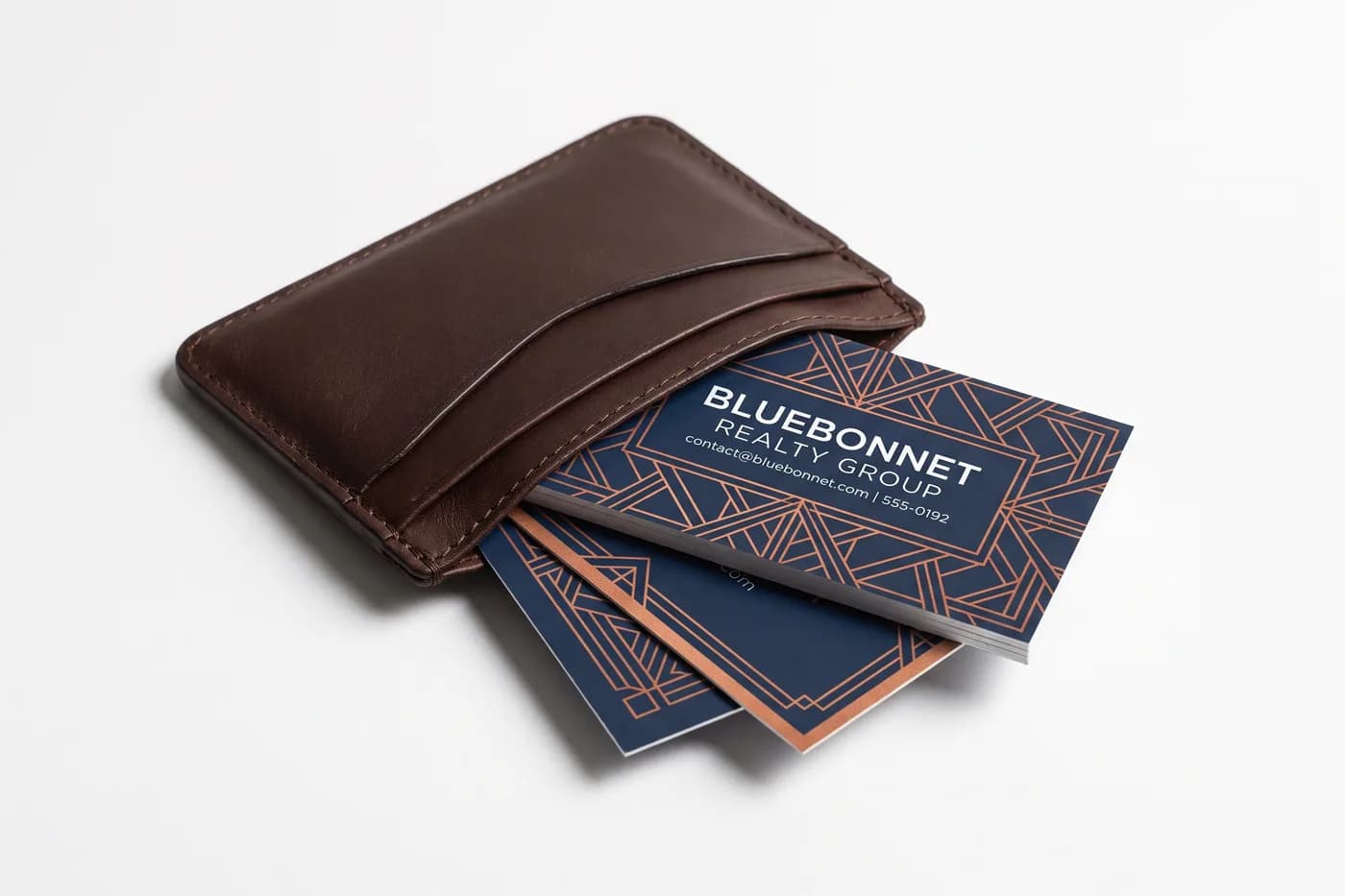 A navy and copper business card for the fictional Bluebonnet Realty Group fanned out of a slim brown leather card wallet on a white surface