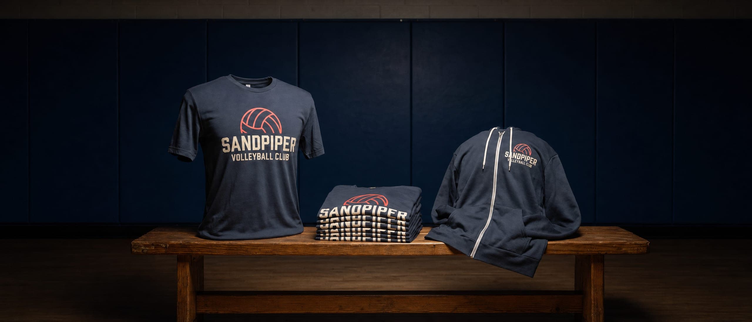 Navy volleyball club printed t-shirt and a folded stack of matching shirts on a wooden bench against a dark wall