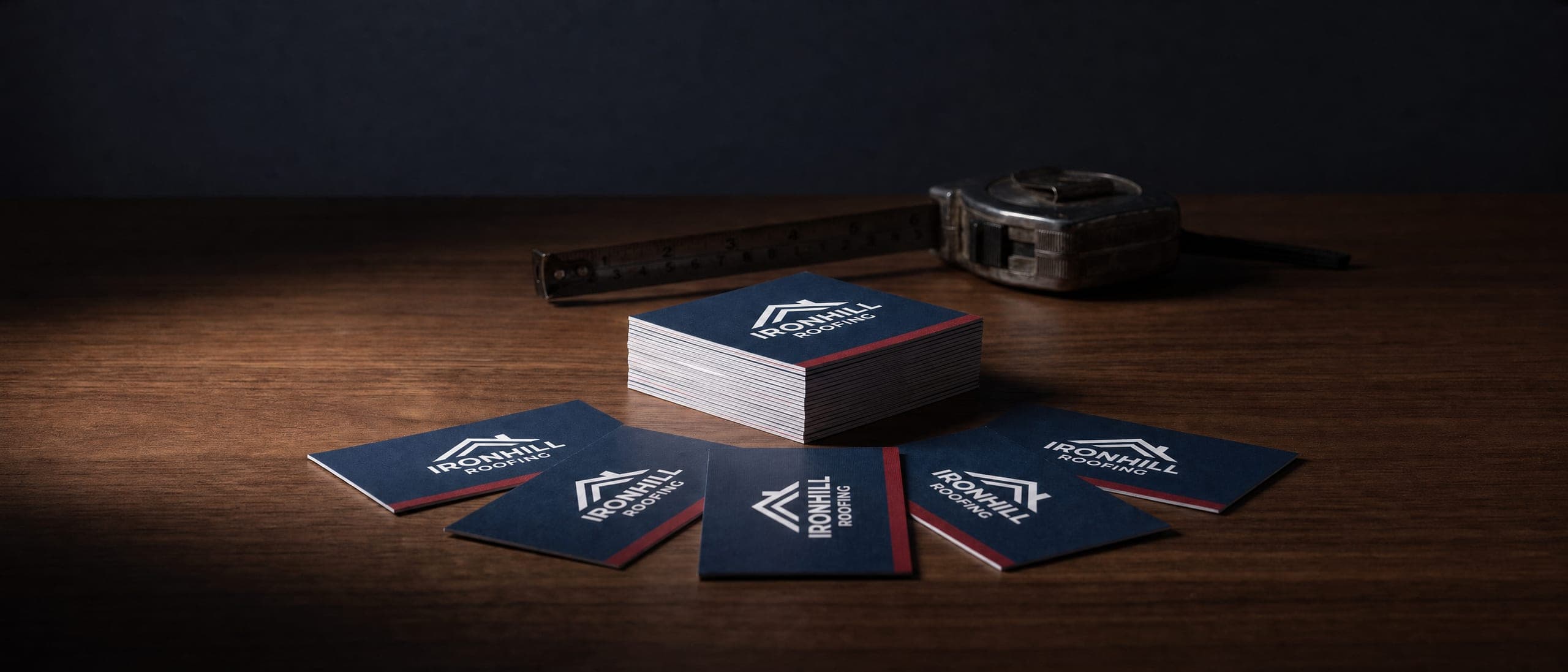 Stack and fan of navy roofing company business cards with red edges on a dark wood desk beside a tape measure