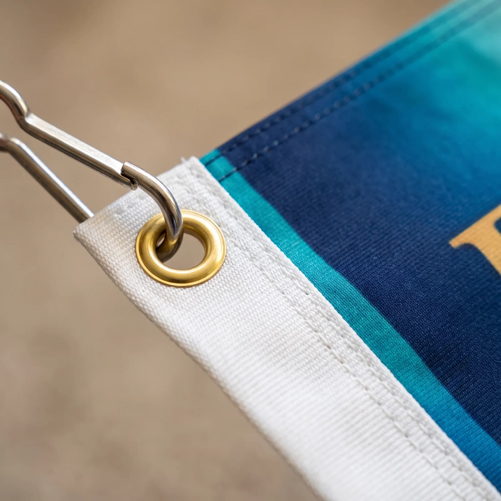 Close up of a stainless snap hook clipped through a brass grommet in the white hem and grommet strip on the hoist edge of a navy and teal printed flag
