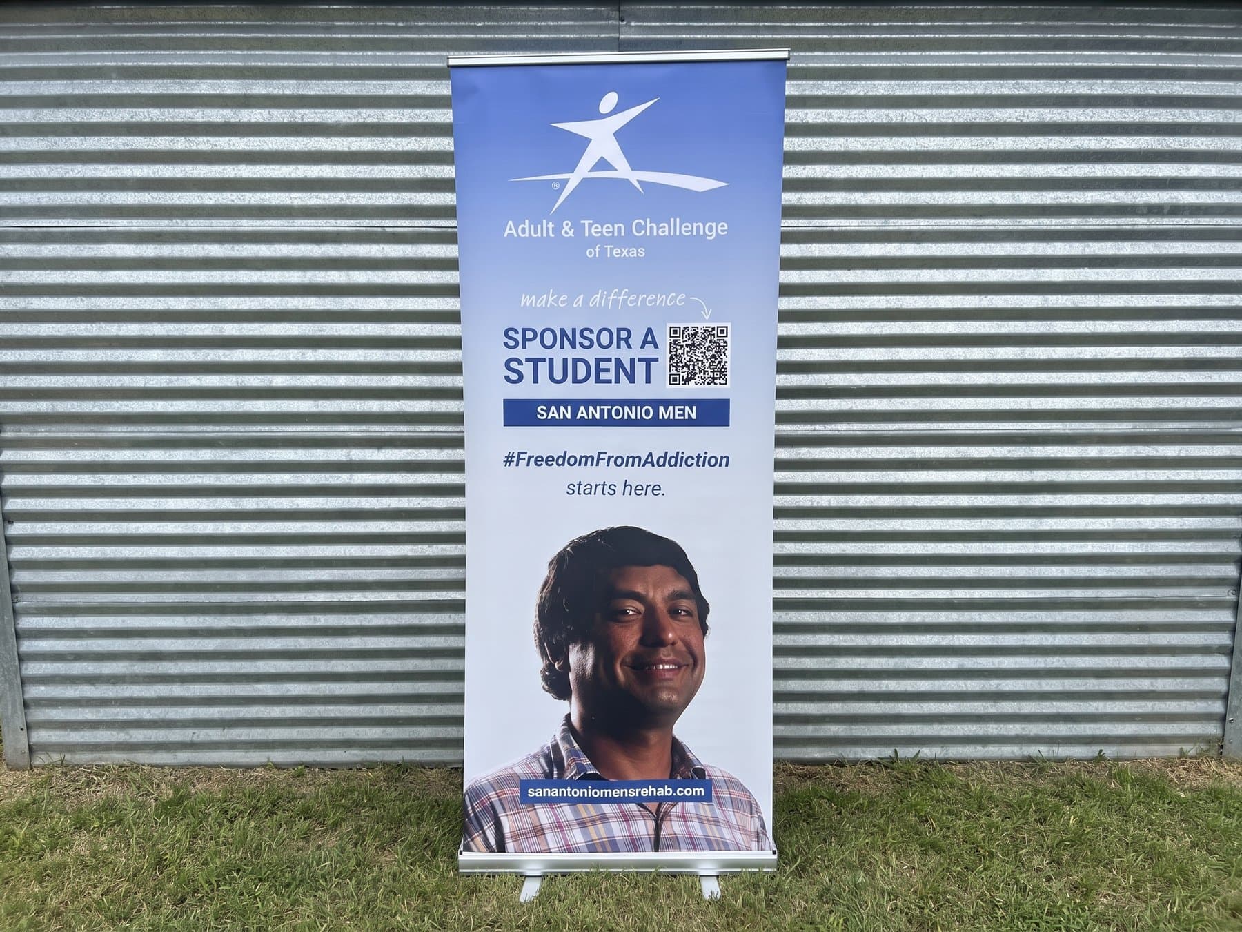 Adult and Teen Challenge of Texas retractable banner standing outdoors against a metal wall, the Sponsor a Student ask with a scannable code beside it and a student's portrait filling the lower half
