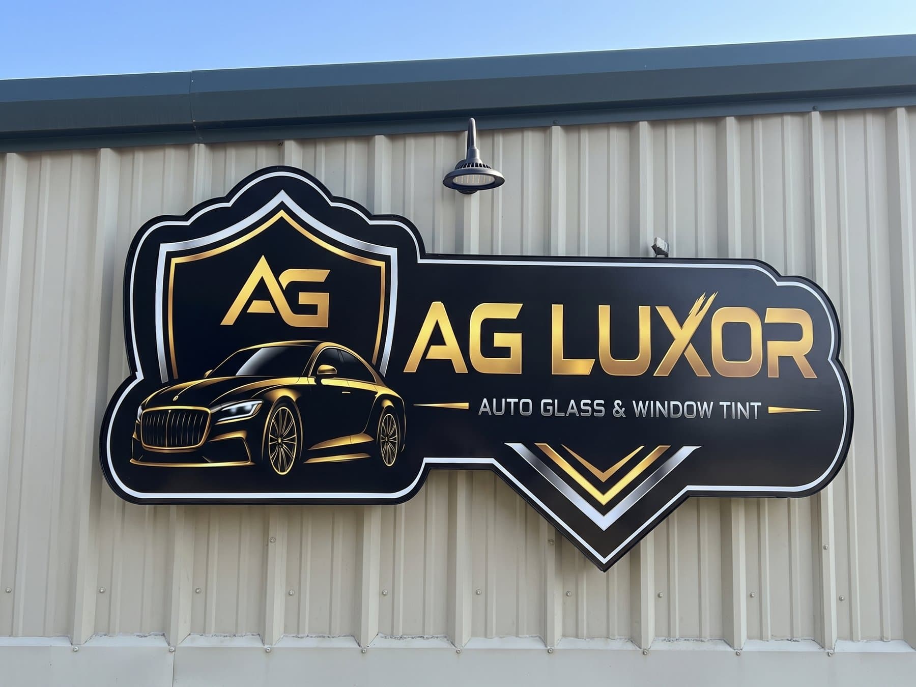 Contour cut AG Luxor Auto Glass and Window Tint sign centered on the corrugated metal wall in sunlight, with the gold shield, the gold car and the chevrons under the service line