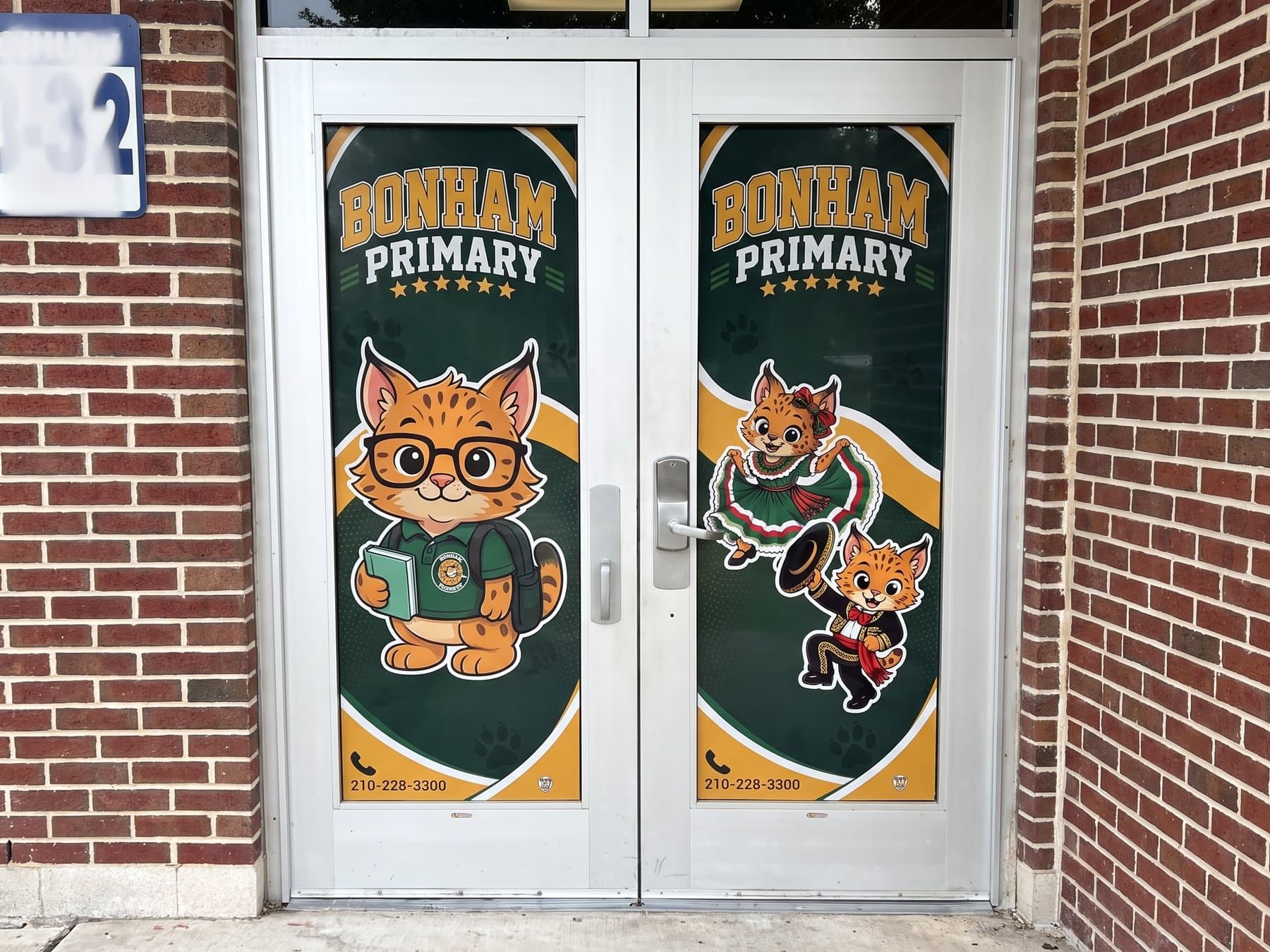 Bonham Primary bobcat mascot graphics on both leaves of the school entry door, each leaf carrying a different pose under the arched school name lockup with the office phone number along the bottom