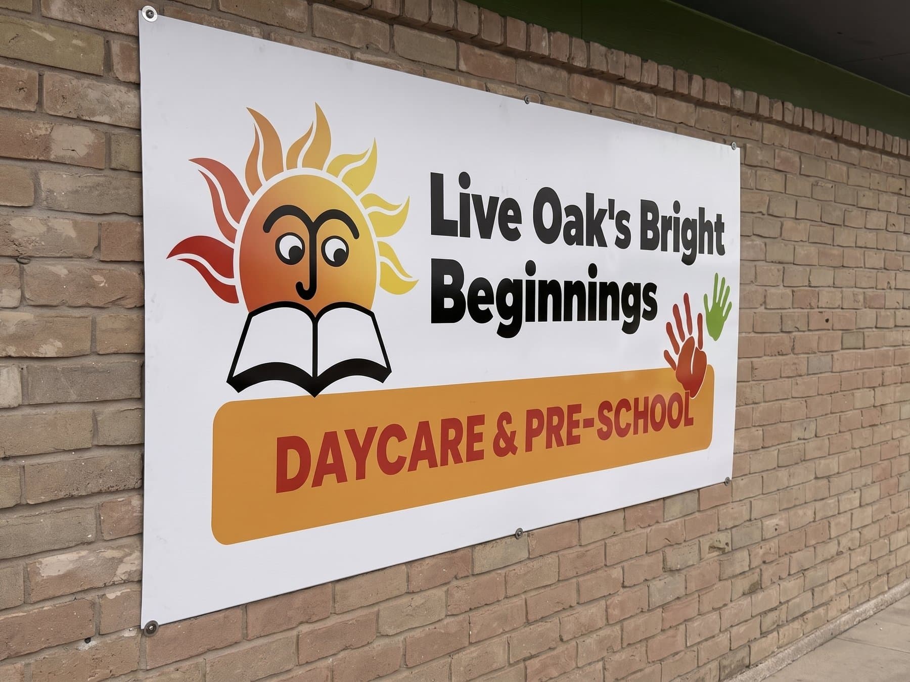 Live Oak's Bright Beginnings wall sign mounted flat on the brick beside the entrance in daylight, showing the sun and open book logo, the two handprints and the orange Daycare and Pre-School bar