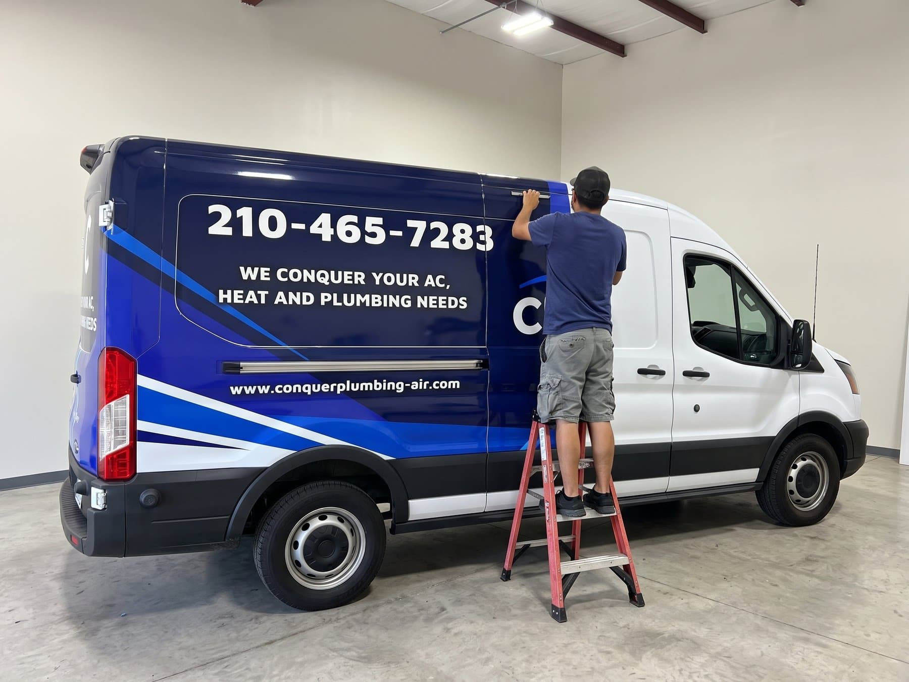 Installer on a step ladder squeegeeing a panel of the Conquer Plumbing and Air van wrap in the shop, the navy and blue field already carrying the phone number in the largest type on the body