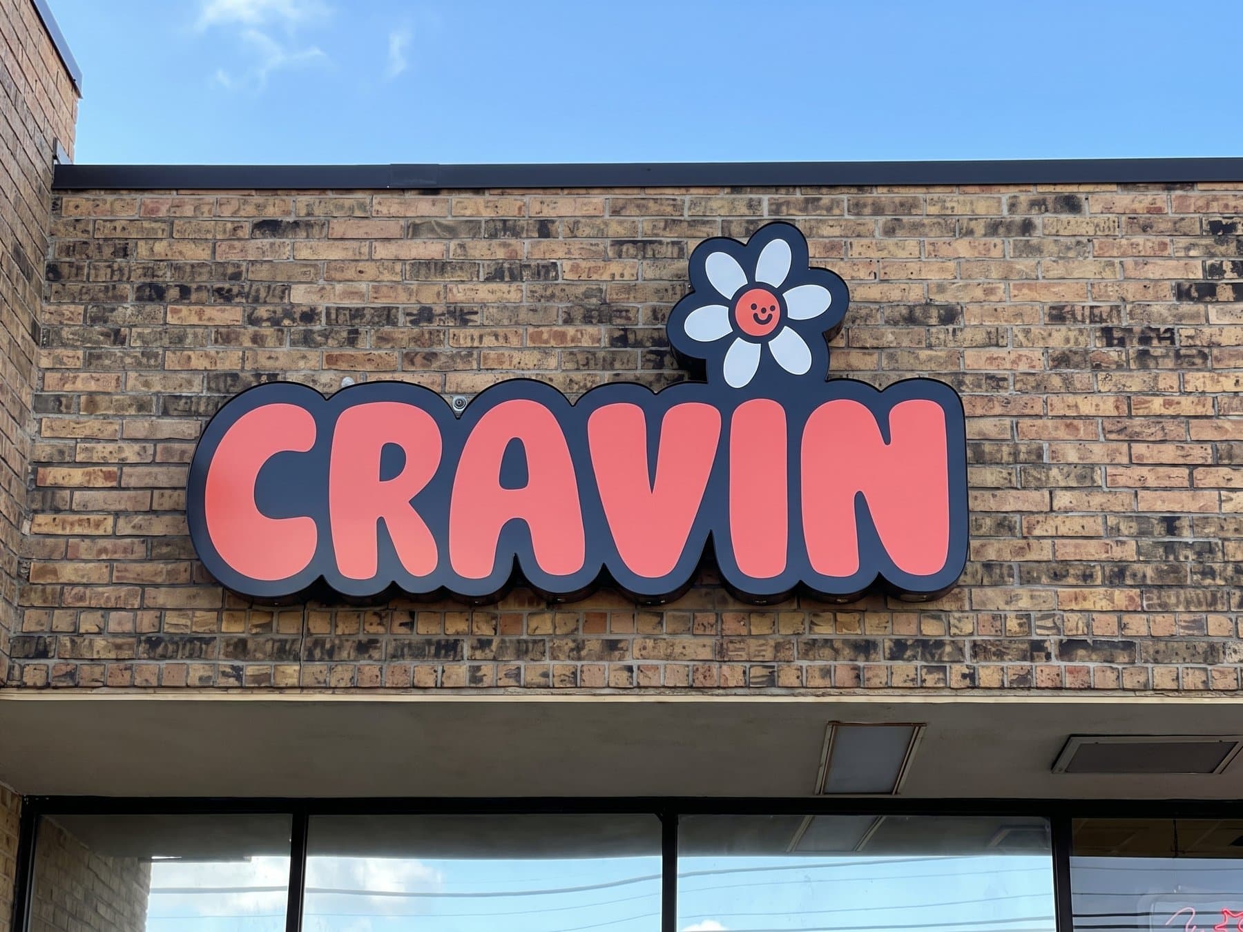 Cravin storefront sign centered on the brick fascia in full sun, coral bubble letters with dark returns and the smiling daisy sitting above the N