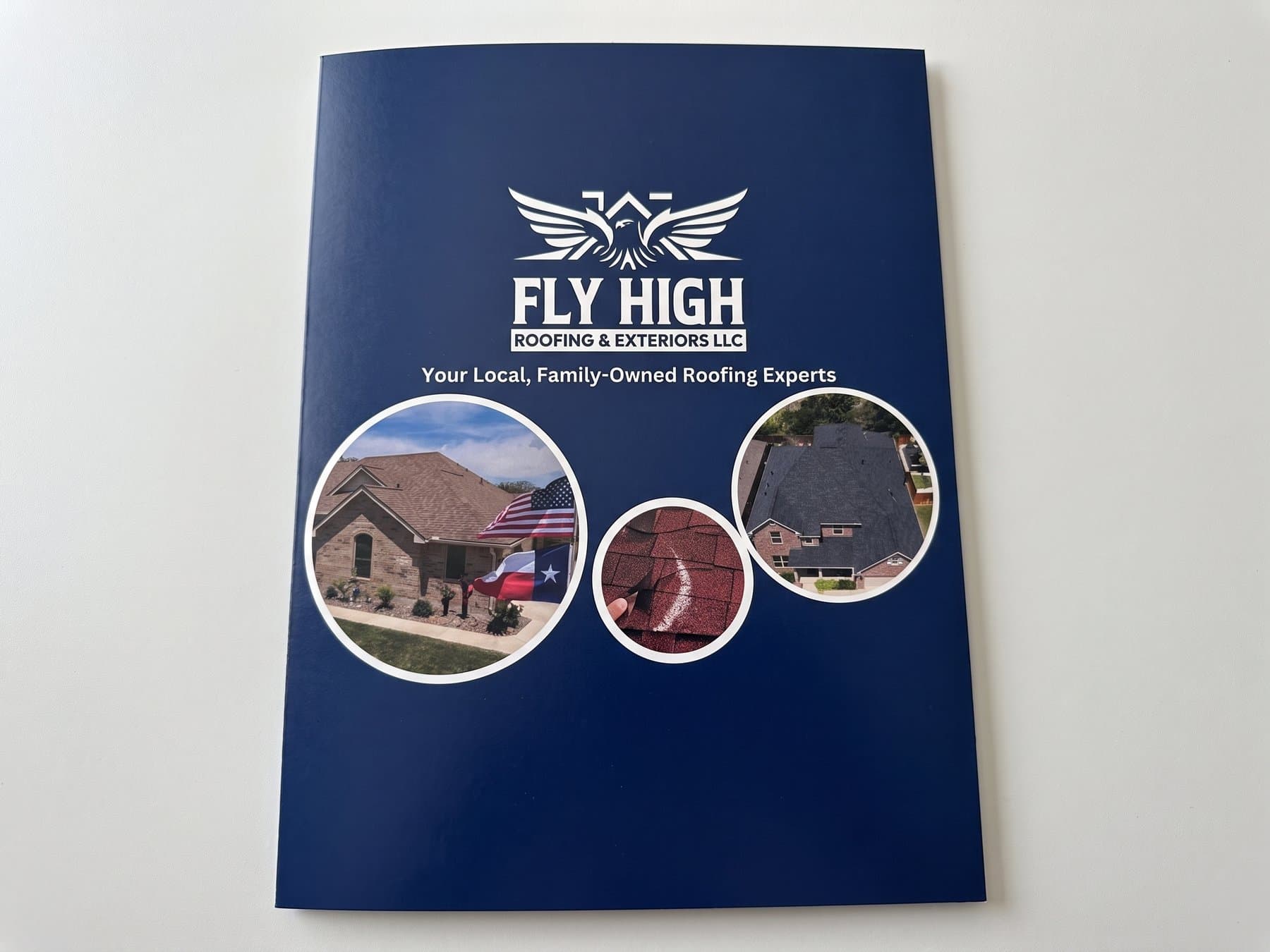 Closed Fly High Roofing presentation folder photographed flat, showing the eagle mark, the family owned line and three job photographs on the navy cover