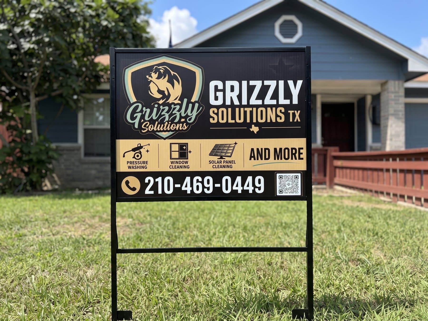 Grizzly Solutions yard sign on its black H-frame stake in a front yard on a sunny day, with the bear mark, three service icons, the phone number and a scannable code