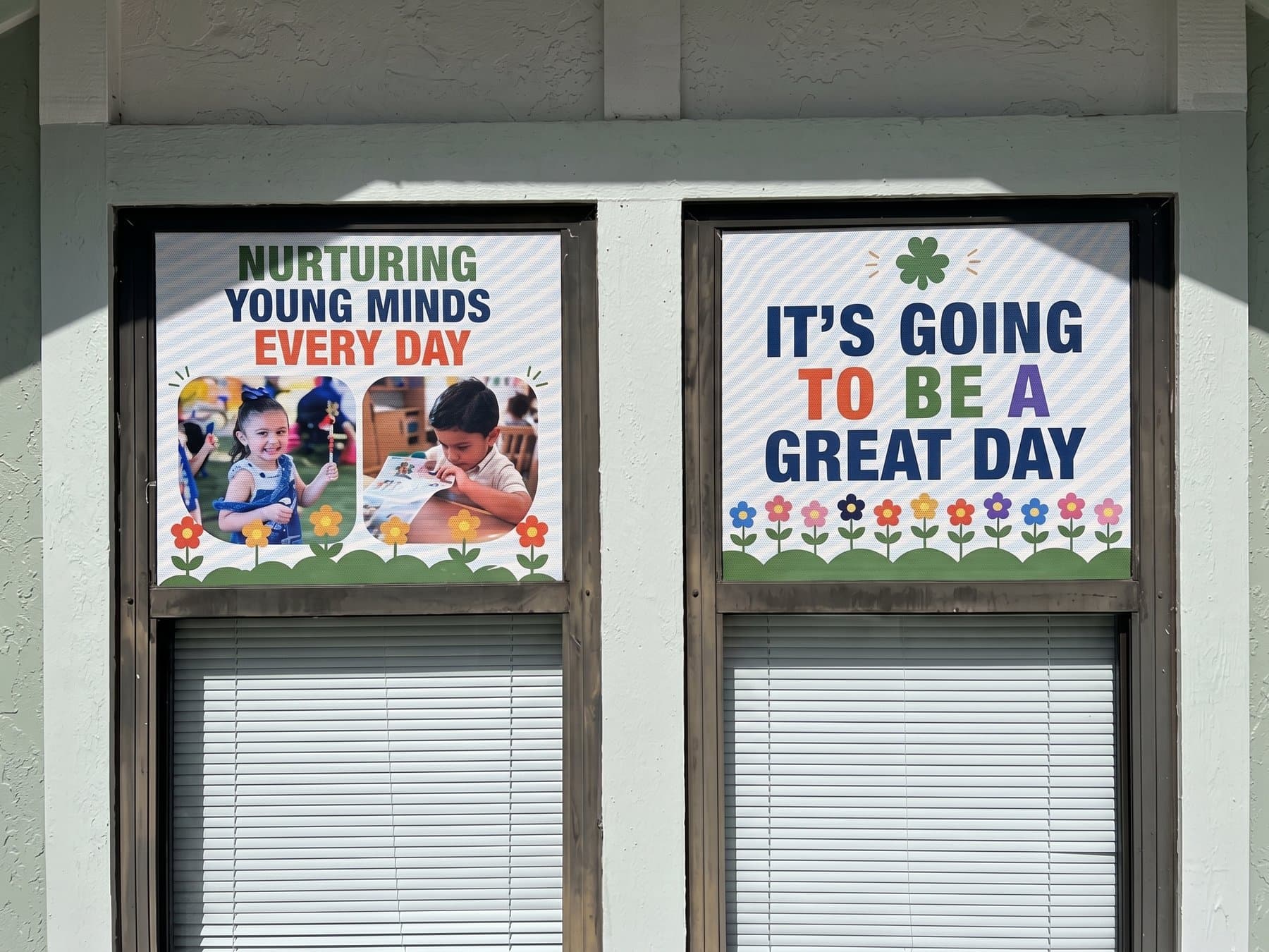 Two upper window sashes on the Growing Minds building carrying perforated panels, one with children's photography under Nurturing Young Minds Every Day and the other reading It's Going To Be A Great Day over a flower border