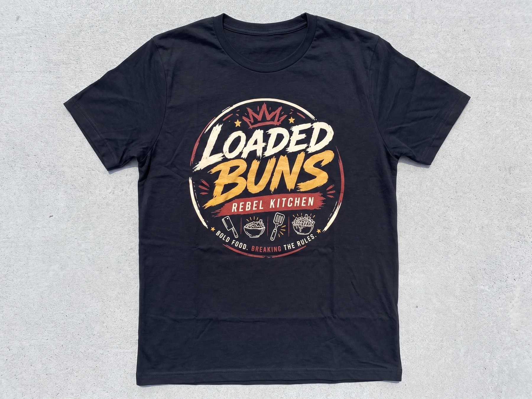 Black Loaded Buns by Rebel Kitchen crew shirt laid flat and centered, showing the full color circular front print with the crown and the utensil icons
