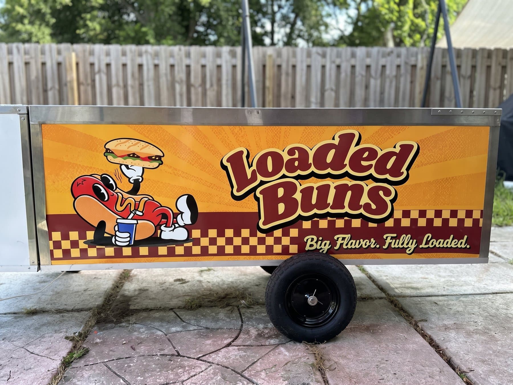 Straight-on view of the Loaded Buns cart side panel outdoors, with the hot dog mascot, the Loaded Buns wordmark, the checker border and the Big Flavor, Fully Loaded tagline