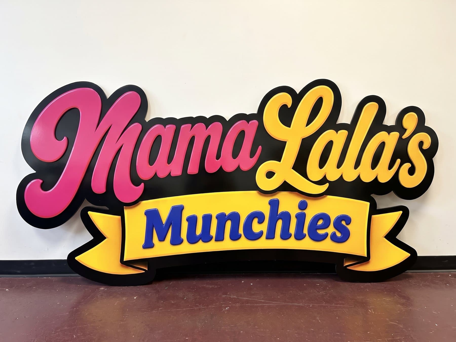 Layered Mama Lala's Munchies sign standing on the shop floor before install, with the pink and yellow scripts raised off the black backer and the Munchies ribbon on its own plane in front