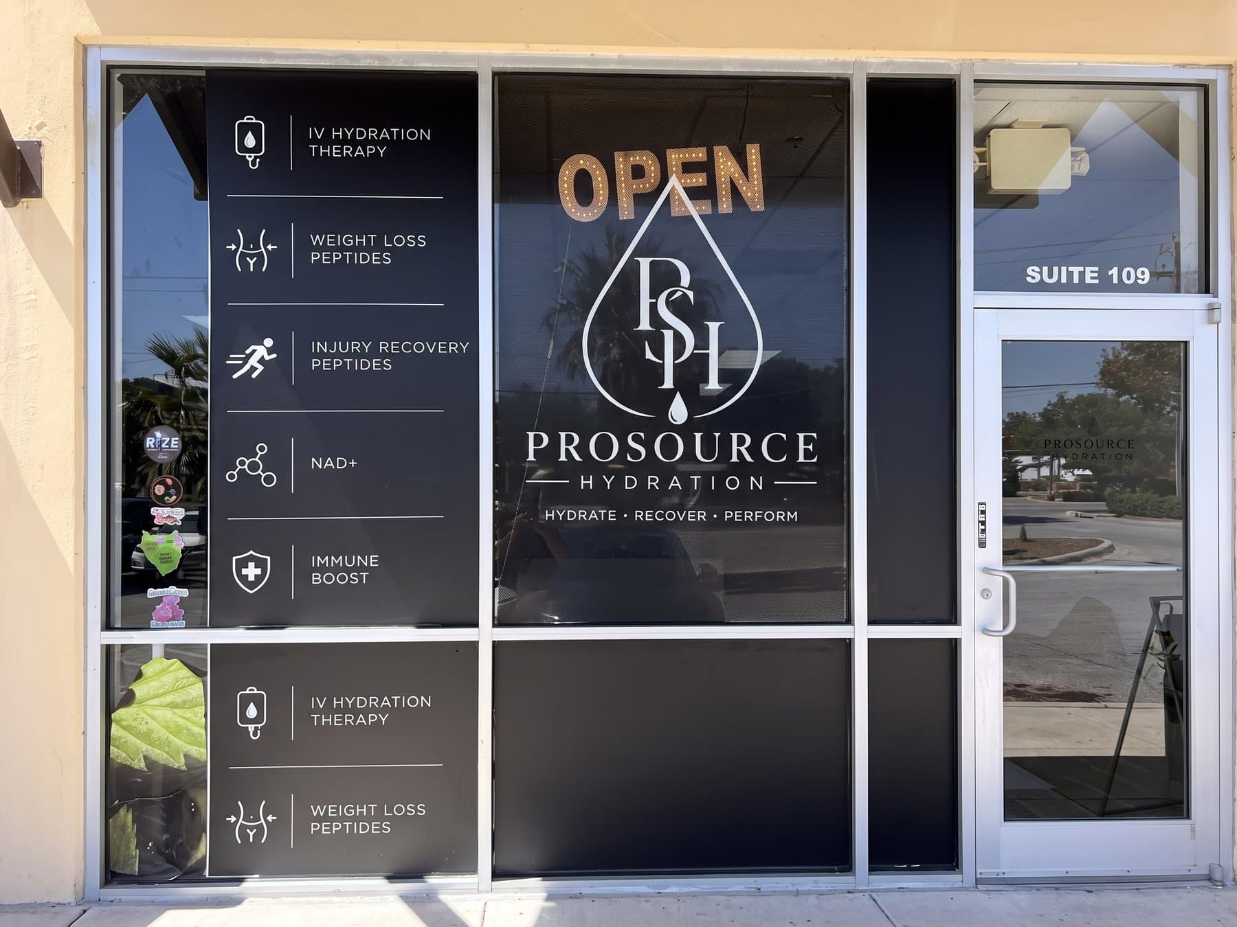 ProSource Hydration storefront under the walkway canopy with every pane covered in black perforated film, the white service menu on the left panes and the drop logo centered beneath a gold OPEN sign
