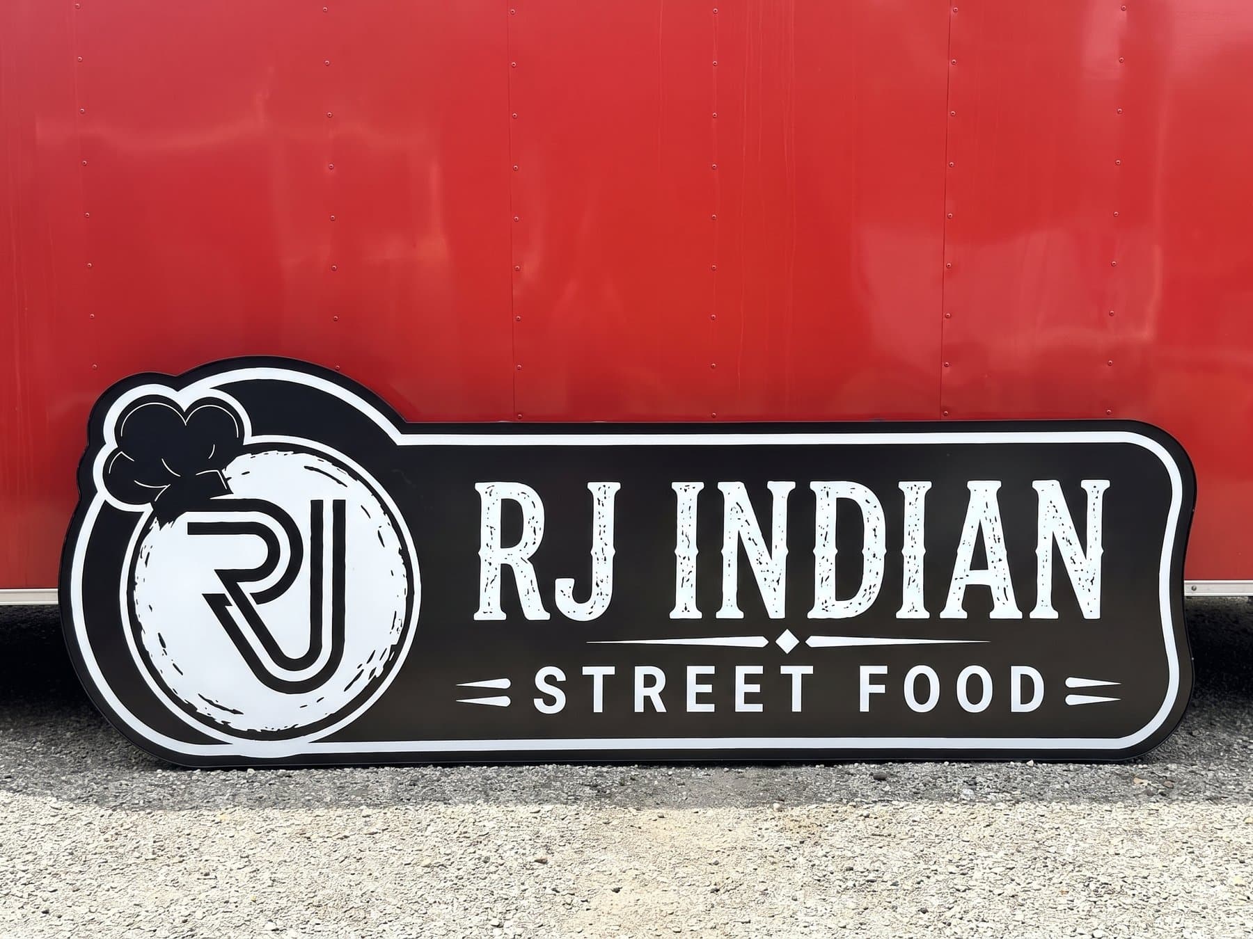 Contour cut RJ Indian Street Food sign propped against the client's red food trailer in daylight, with the chef hat monogram and the distressed white lettering on the black face