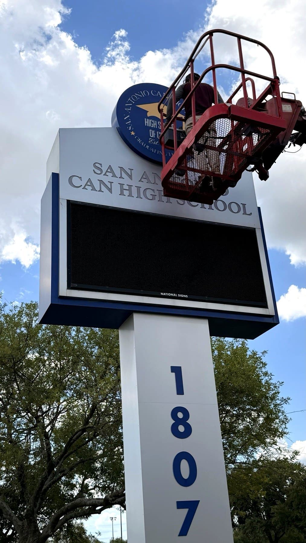 Installer in a crane bucket mounting the round school seal on top of a white and blue pylon sign with dimensional letters and a message center