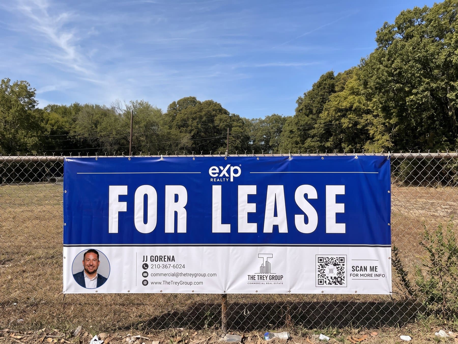 For Lease banner zip-tied along a chain-link fence at a vacant lot, FOR LEASE in white on brokerage blue with the agent's photo, contact details and a Scan Me code in the white band below