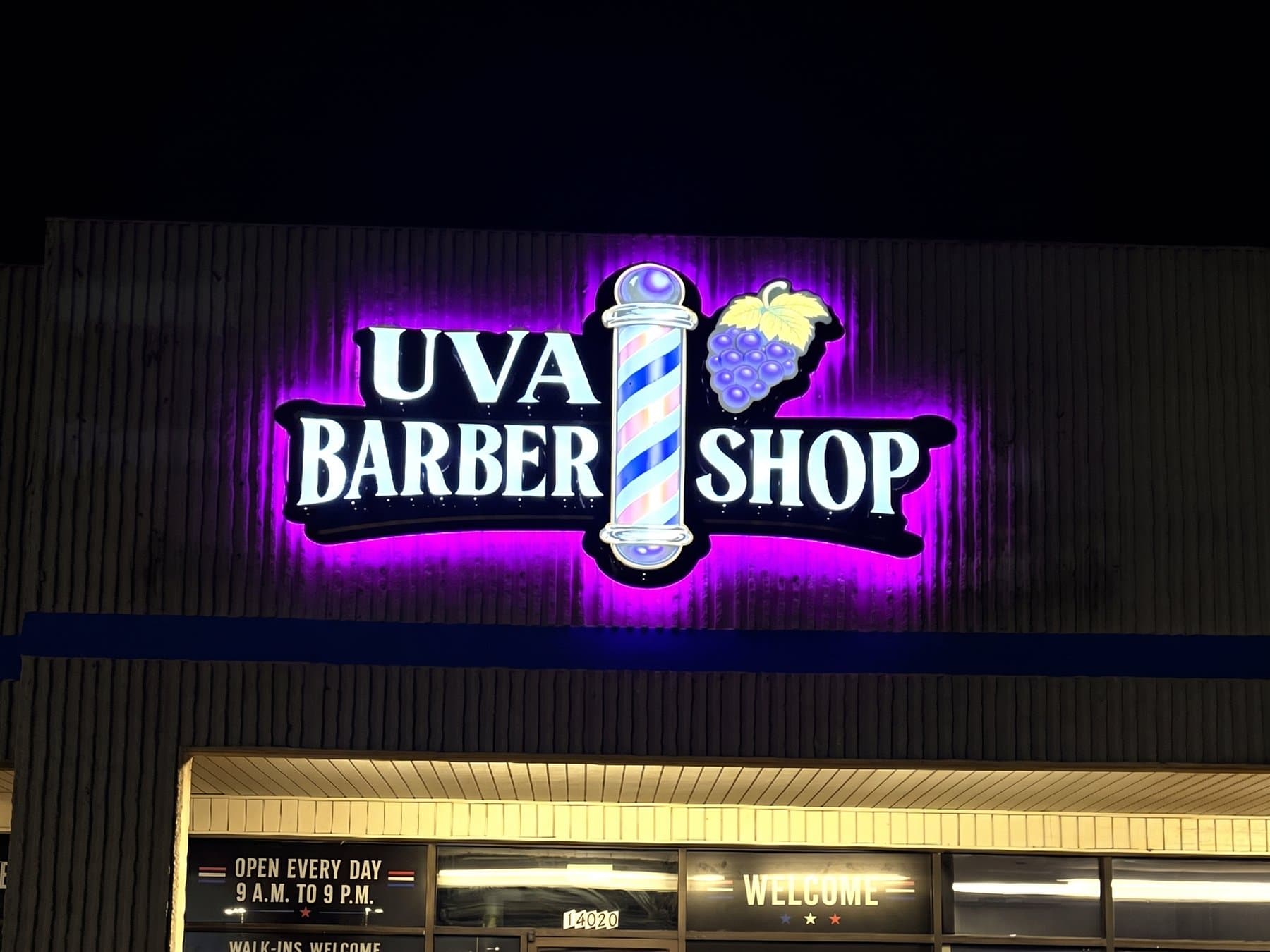 UVA Barber Shop sign glowing on the storefront fascia at night, the halo lighting throwing purple across the metal while the barber pole, the grapes and the lettering stay readable