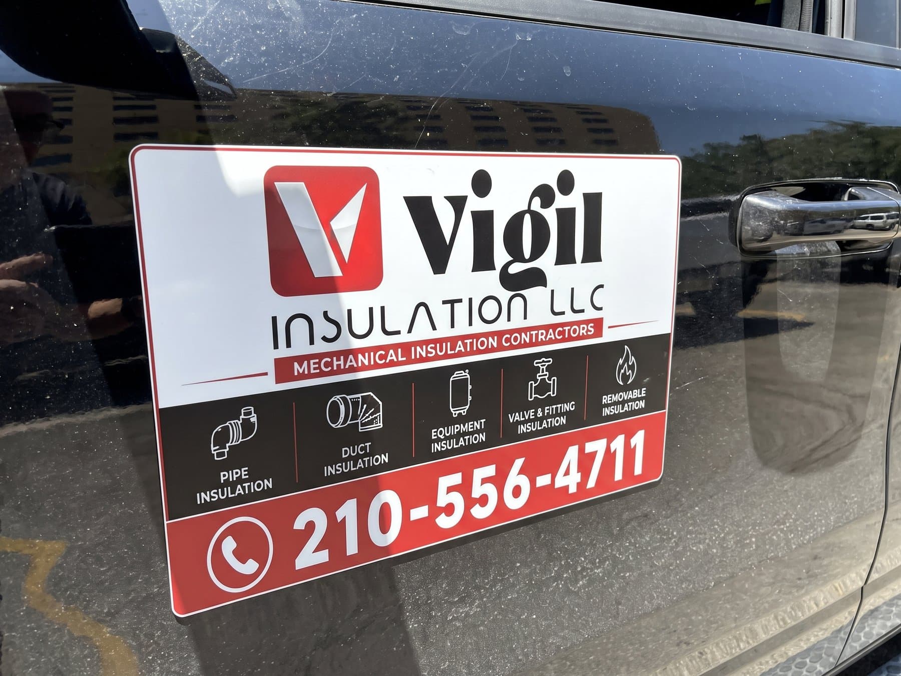 Vigil Insulation door magnet on the driver's door of a dark pickup, with the V mark, the mechanical insulation contractors line, a row of service icons and the phone number in a red band